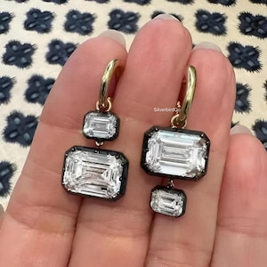 May include: A pair of drop earrings featuring gold hoops and two emerald-cut clear stones set in dark metal frames. One earring has a larger stone on the bottom, while the other has a larger stone on the top. The text SilverbirdCo is visible.