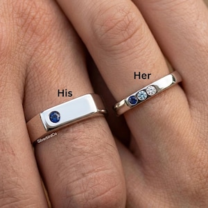 Può includere: Due anelli d'argento, uno con una singola pietra blu e la parola "His", l'altro con tre pietre preziose e la parola "Her". Gli anelli sono su dita, con uno sfondo sfocato.