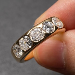 Peut inclure: Une bague en or avec cinq diamants ronds sertis en ligne. La bague est tenue par une main, mettant en valeur son anneau en or poli. Les diamants scintillent, soulignant le design élégant de la bague. Un bijou classique.