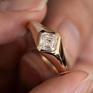 Puede incluir: Un anillo de oro con un diamante de talla cuadrada. El diamante está engastado en un bisel, y la banda es lisa y pulida. El anillo se sostiene entre dos dedos.
