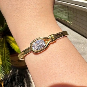 May include: Gold-toned bracelet featuring a large, clear, oval-shaped stone. The bracelet has a unique design with a knot detail and a sleek, polished finish. A classic piece of jewelry.