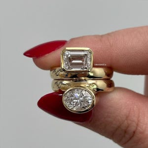 Puede incluir: Tres anillos de oro con piedras preciosas de diamantes. El anillo superior presenta un diamante talla esmeralda, el anillo central tiene una banda texturizada y el anillo inferior muestra un diamante talla ovalada. Los anillos se sostienen sobre un fondo claro.