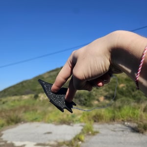 Op de afbeelding: Een zwart, getextureerd object met een puntige vorm, gehouden tegen een helderblauwe lucht en een groene heuvel. Het object lijkt een klein, mogelijk decoratief item te zijn, met een dun blauw touwtje eraan.
