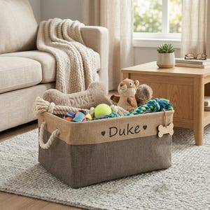 Puede incluir: Una cesta rectangular para guardar juguetes para perros con asas de cuerda, llena de juguetes para perros. La cesta es marrón y beige con el nombre "Duke" impreso en la parte delantera. Una etiqueta en forma de hueso cuelga del lateral.