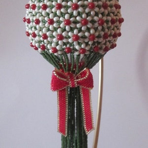 Puede incluir: Un adorno navideño de cuentas con un diseño floral verde y blanco y un lazo rojo. El adorno está colgado de un soporte dorado.