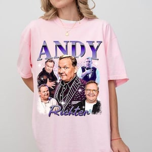 May include: A light pink t-shirt featuring a graphic of Andy Richter in various poses, with the name "ANDY" in purple block letters above and "Richter" in cursive below. The design has a vintage aesthetic.