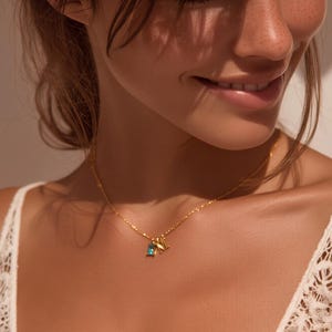 Puede incluir: Un collar de cadena en tono dorado con dos pequeños dijes: una piedra preciosa rectangular de color azul brillante y un colgante con la letra M en tono dorado. El collar descansa sobre la clavícula con un diseño de eslabones finos.