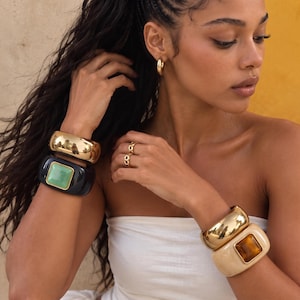 May include: A set of four wide cuff bracelets and two rings. One wrist features a polished gold-tone cuff and a black cuff with a square green stone. The other wrist has a gold-tone cuff and a beige cuff with a square amber-colored stone.