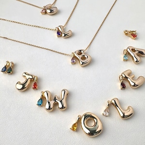 May include: Gold-coloured initial necklaces and charms with colourful gemstone accents. The letters are in a playful, rounded font. Some necklaces are displayed on a white surface. The gemstones are teardrop-shaped and in various colours.