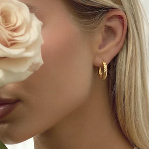 May include: A close-up shot of a person's ear adorned with a gold hoop earring. The earring features a textured, ridged pattern. The person has blonde hair and fair skin. A white rose is partially visible.