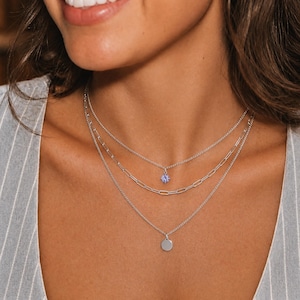 May include: A layered silver-tone necklace set consisting of three distinct chains. The top chain features a small, round, light purple faceted stone pendant. The middle chain is a paperclip link style. The bottom chain holds a small, flat, round silver disc.