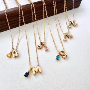May include: A collection of gold-toned necklaces, each featuring a polished letter pendant paired with a small, teardrop-shaped gemstone charm. The letters A, M, J, S, K, and R are displayed, accompanied by stones in yellow, dark blue, pink, teal, clear, and light blue.