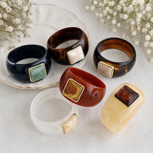 Peut inclure: Un ensemble de six bracelets joncs larges en résine de différentes couleurs, notamment noir, brun foncé, écaille de tortue, brun rougeâtre, blanc et crème. Chaque bracelet présente une incrustation carrée sertie d'un fin bord en métal doré.