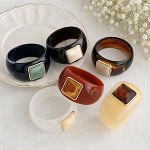 May include: A collection of five wide resin bangle bracelets in various colors including black, dark brown, reddish-brown, and cream. Each bracelet features a central square-shaped inlay framed in gold-tone metal, showcasing unique marbled patterns.