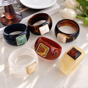 Op de afbeelding: Zes dikke harsringen in diverse kleuren, waaronder zwart, bruin schildpad, donkerrood en crème. Elke ring heeft een gladde, ronde band en een centrale vierkante steen of inleg geplaatst in een dunne, goudkleurige metalen rand.