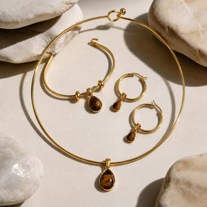 May include: A gold-toned jewelry set consisting of a rigid circular choker necklace, a hook-style bracelet, and two hoop earrings. Each piece features a dangling, teardrop-shaped tiger's eye gemstone pendant with a smooth, polished brown finish.
