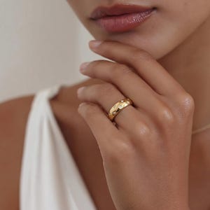 May include: A gold ring with a rounded shape, set with small, clear gemstones. The ring is displayed on a finger, highlighting its polished finish and delicate features. The ring's design is understated yet stylish.