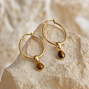 May include: A pair of gold-toned hoop earrings, each featuring a single teardrop-shaped tiger's eye gemstone pendant suspended from the bottom of the hoop. The earrings have a polished metallic finish and a secure hinged closure mechanism.