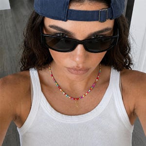 May include: A white ribbed vest top worn with a multicoloured beaded necklace featuring a gold-toned cursive letter charm. The outfit includes dark-tinted rectangular sunglasses and a navy blue baseball cap worn backwards with an adjustable metal buckle.