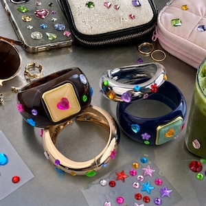 Pode incluir: Três pulseiras grossas com adornos de strass coloridos. Uma é castanho-escura com uma placa quadrada central, uma é de metal prateado com gemas em forma de flores e corações, e uma é um bracelete de metal dourado com cristais espalhados.