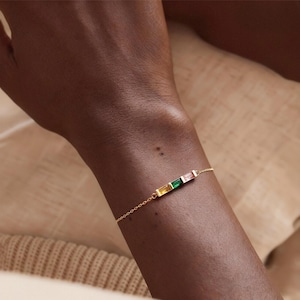 May include: A gold bracelet with a fine chain and a bar featuring four rectangular gemstones in yellow, green, pink, and a darker pink colour. The bracelet is worn on a wrist.