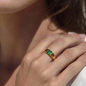 Puede incluir: Anillo de oro con una piedra verde rectangular con talla esmeralda. El anillo tiene una banda lisa y pulida y un engaste biselado. El anillo se muestra en un dedo.