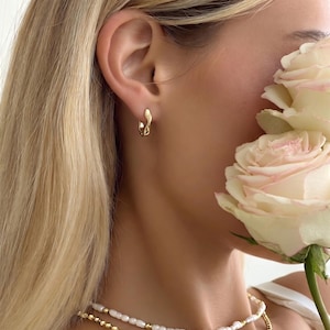 May include: A close-up shot of a person's ear adorned with a small, gold hoop earring. The earring features a distinctive, fluid design. A pearl necklace is also visible. A large, white rose is partially in view.