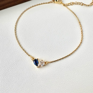 Peut inclure: Un bracelet doré délicat avec deux pierres précieuses en forme de goutte. Une pierre est bleu foncé, l'autre est transparente. Le bracelet a une fine chaîne et une fermeture à fermoir homard.