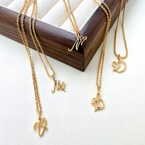 Op de afbeelding: Goudkleurige initiaal kettingen op een witte ondergrond. Elke ketting heeft een fijne ketting en een hanger met een cursieve letter. De letters zijn V, M, M, D en A. De kettingen liggen in de buurt van een houten doos.