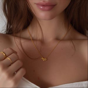 May include: A gold-colored necklace with the letters "mia" hanging from a delicate chain. The image also shows two gold rings on a finger.