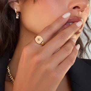 May include: A gold signet ring featuring a starburst design and a small gemstone. The ring is worn on a finger. The image also shows a gold chain necklace and gold hoop earrings.
