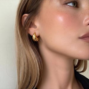 May include: A close-up of a gold-coloured teardrop-shaped earring. The earring has a smooth, polished surface and a rounded, hollow design. The earring is worn on the ear of a person with blonde hair.