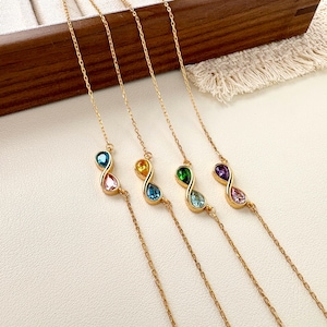 May include: Four gold-toned chain bracelets, each featuring an infinity symbol centrepiece set with two pear-shaped gemstones. The stones include combinations of blue, pink, yellow, green, and purple, arranged in various colour pairings on each bracelet.