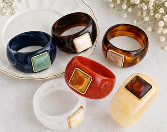 May include: A collection of six chunky resin bangle bracelets in various colors including dark blue, black, brown tortoiseshell, red, white, and cream. Each bracelet features a square-shaped inset stone with a gold-toned metal border.