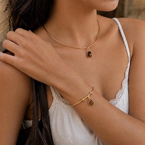 May include: A gold-toned metal choker necklace featuring a single teardrop-shaped brown gemstone pendant, paired with a matching gold-toned metal bangle bracelet that also includes a dangling teardrop-shaped brown gemstone charm.