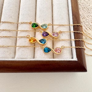 May include: Four gold-tone necklaces featuring infinity-shaped pendants. Each pendant is set with two pear-cut gemstones in various colours, including green, light blue, purple, pink, yellow, and dark blue, attached to a delicate gold-tone chain.