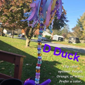 May include: A stroller fan accessory featuring a vertical rod adorned with multicolored beads in shades of purple, orange, blue, and white, topped with a cluster of hanging ribbons. The text D Duck and Clip color options: Purple, Orange, or White. Prefer a color, add to personalization. is visible.