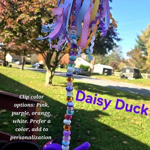 May include: A decorative accessory featuring a vertical strand of multi-colored beads attached to a purple clip. The top is adorned with a cluster of various ribbons in shades of pink, purple, yellow, and blue. Text reads: Daisy Duck. Clip color options: Pink, purple, orange, white. Prefer a color, add to personalization.