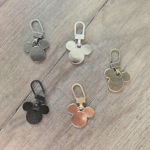 Może przedstawiać: Pięć metalowych uchwytów do zamków błyskawicznych w różnych wykończeniach: srebrnym, czarnym, złotym i brązowym. Każdy uchwyt ma wzór głowy Myszki Miki i metalowe zapięcie do mocowania. Uchwyty są ułożone na drewnianej powierzchni.