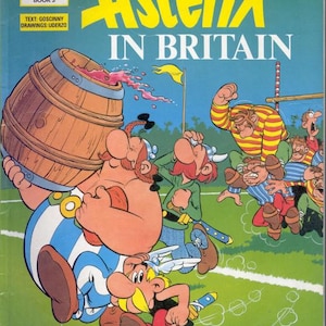 Pode incluir: Capa da banda desenhada "Astérix na Grã-Bretanha". A capa apresenta uma ilustração de desenho animado de personagens da série Astérix. O título está em letras amarelas grandes. Um grande barril de madeira está sendo derrubado, derramando líquido vermelho.