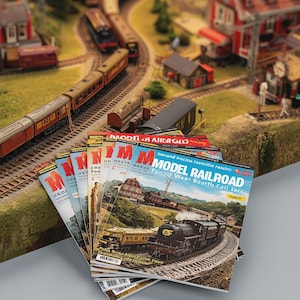 May include: A stack of model railroad magazines with colorful covers, featuring images of trains and landscapes. The magazines are placed in front of a detailed model train layout with miniature buildings, trees, and tracks.