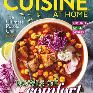 May include: The cover of "Cuisine at Home" magazine features a vibrant bowl of posole chili, garnished with red cabbage, radishes, and lime wedges. The magazine title is in large yellow letters, with the words "Bowls of Comfort" prominently displayed.