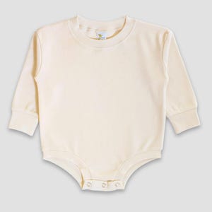 Pode incluir: Body de bebé creme com mangas compridas e gola redonda. A peça única tem fechos de pressão na parte inferior para facilitar a troca. O tecido parece macio e confortável, adequado para bebés.