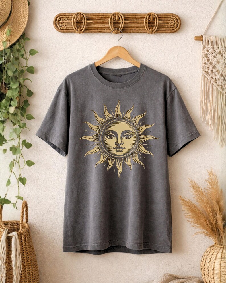 Charcoal gray celestial sun face t-shirt hanging on a wooden hanger, showing a vintage boho astrology design in a clean product display