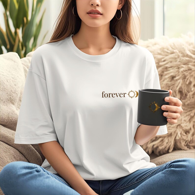 Woman sitting on a couch wearing an off-white tee with small "forever" sun and moon chest print, with a black cup in hand with sun&moon print. Cozy lifestyle photo.