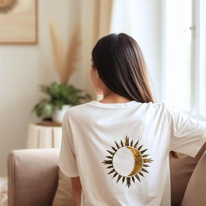 Back view of a white t-shirt with large gold sun and crescent moon graphic, casual indoor lifestyle setting.