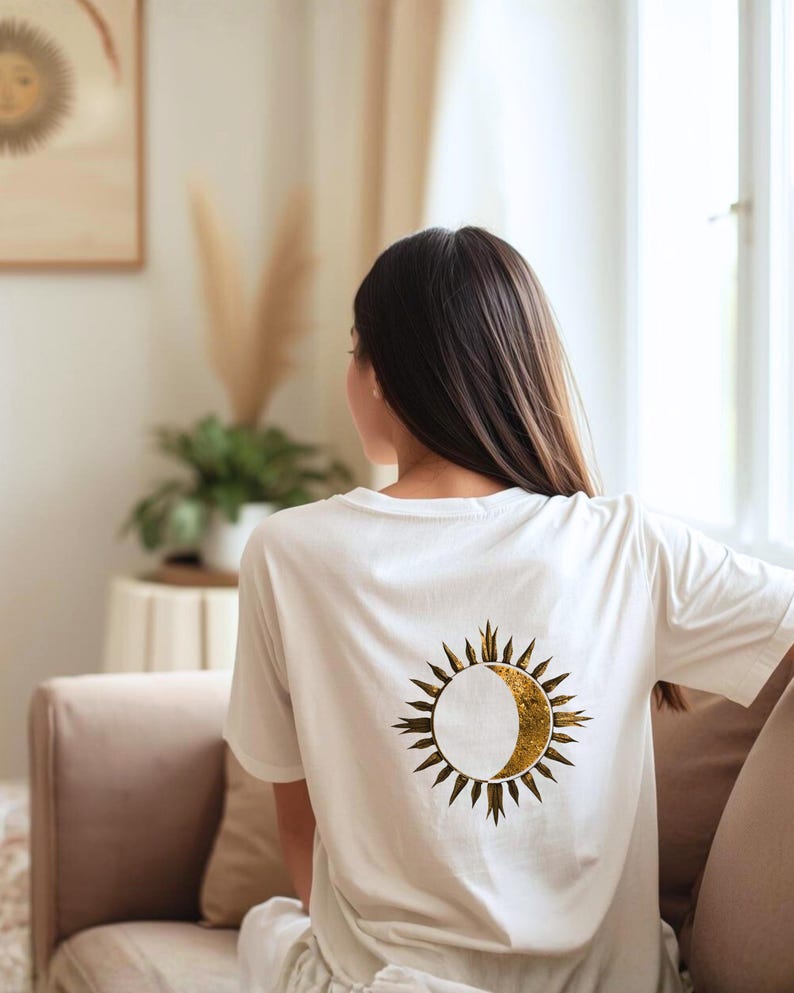 Back view of a white t-shirt with large gold sun and crescent moon graphic, casual indoor lifestyle setting.