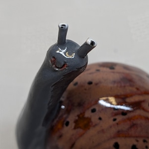 May include: A ceramic snail ornament with a glossy finish. The snail's body is dark grey, and the shell is brown with black spots. The snail has two antennae with silver tips and a smiling face. The ornament is a decorative item.