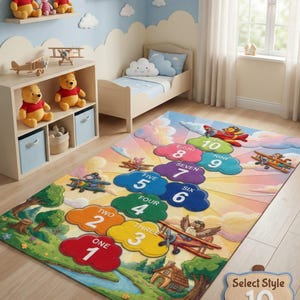 May include: A colorful children's rug with a whimsical design featuring numbers one through ten in cloud shapes. The rug has a landscape scene with airplanes and cartoon characters. The rug is in a child's bedroom with stuffed animals and a bed.