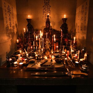 May include: A dark wooden altar table illuminated by numerous lit candles, featuring a central dark statue, various small figurines, bowls, and metal blades. The stone walls behind the altar display vertical columns of black calligraphic text.